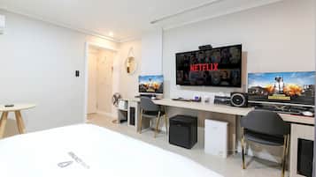 Une chambre d’hôtel moderne équipée d’un grand lit, d’un bureau avec un ordinateur, d’une télévision diffusant une série Netflix et d’un espace dédié aux jeux vidéo.