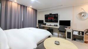 Une chambre d’hôtel comprenant un lit, un bureau avec deux écrans, un téléviseur diffusant Netflix, une table ronde et un petit meuble de rangement.