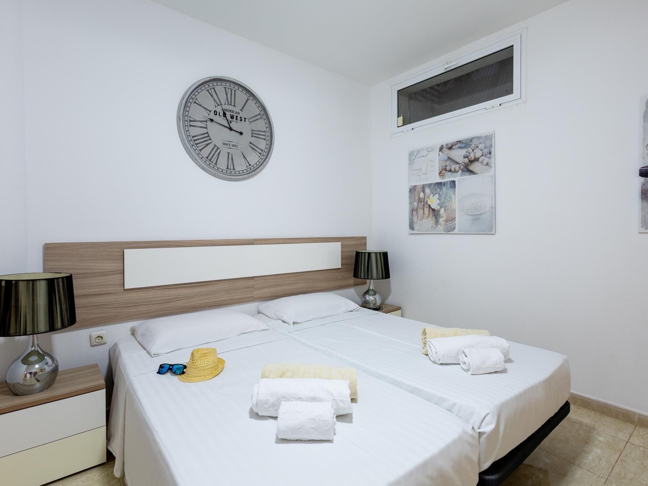 A hotel room with a bed, two bedside tables, a wall clock, and a window.