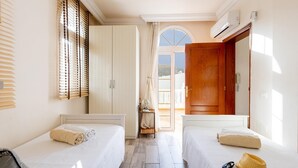 Ein Hotelzimmer mit zwei Betten, einer Holztür, einem Fenster mit Jalousien und Blick nach draußen.