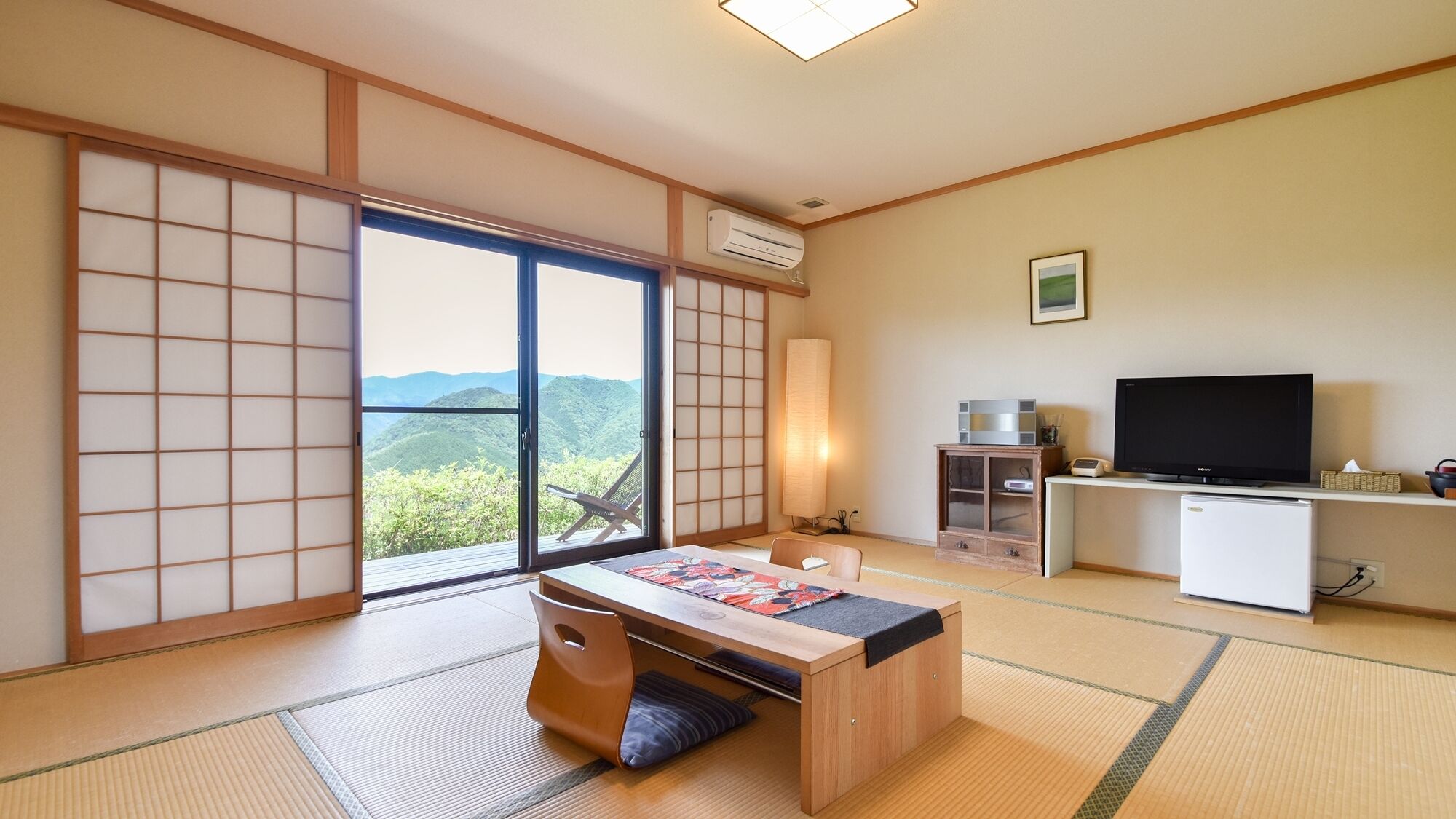 Ein traditioneller japanischer Raum mit Tatami-Boden, einem niedrigen Holztisch, einem Fernseher und einer Schiebetür mit Blick auf Berge.