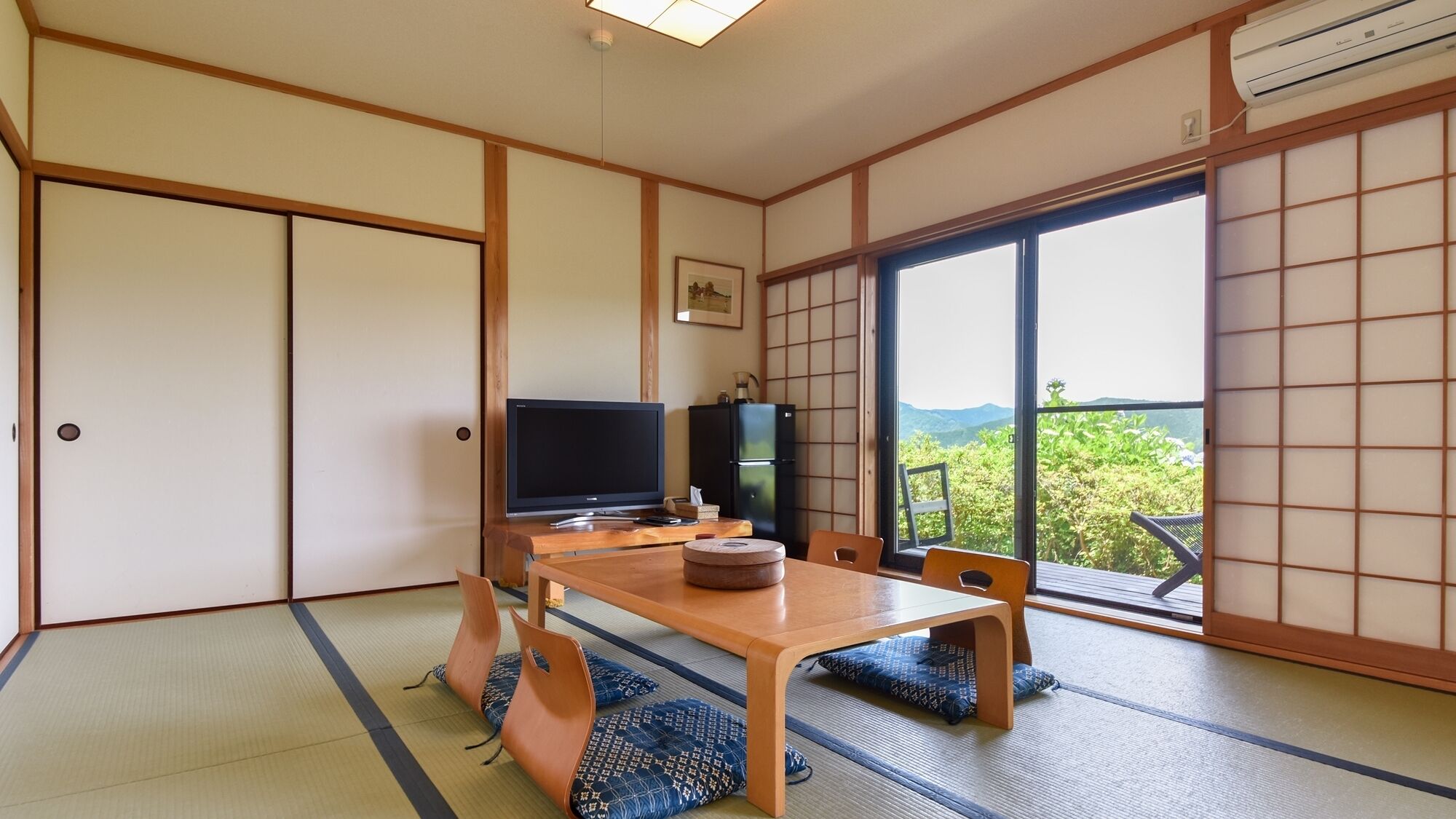 Ein traditioneller japanischer Raum mit Tatami-Böden, einem Fernseher, einem Kühlschrank und Blick auf Grün hinter großen Fenstern.