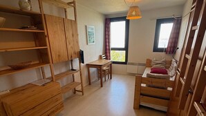 A wooden room with a bed, a table, and a chair. There is a window with curtains, a hanging light, and a radiator. - Studio for 3, slopeside in Les Adrets (Les Adrets-Prapoutel)