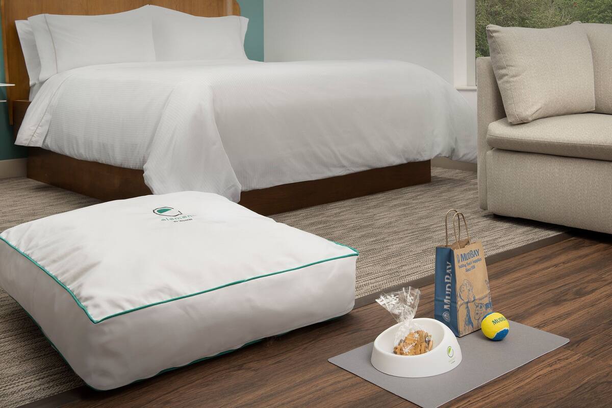 A neatly made bed with white bedding, a white pillow with teal trim, a grey yoga mat, a bowl of snacks, and a MUDPAVY bag on the floor.