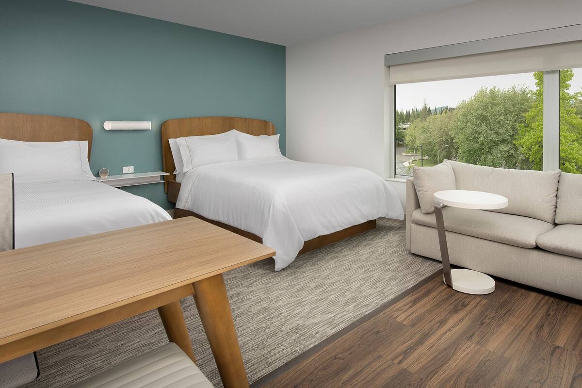 A hotel room with two beds, a sofa, a small table, and a large window offering a view of greenery.