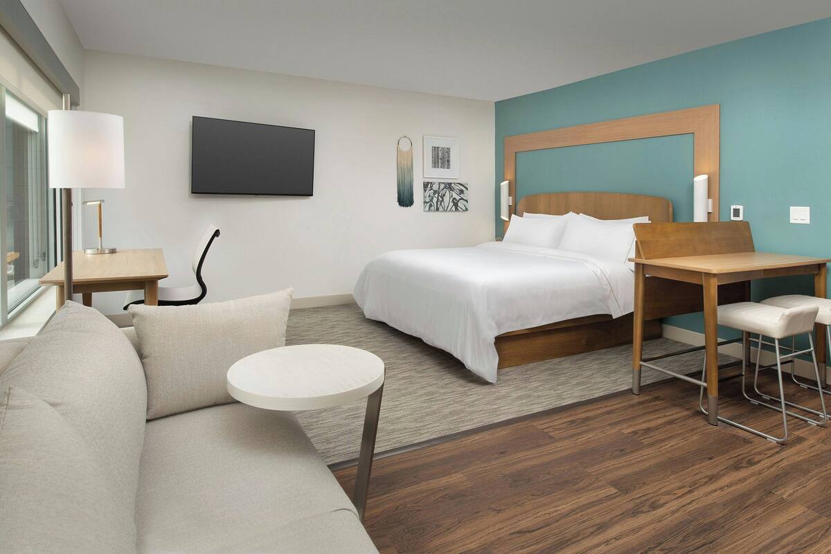 A modern hotel room with a bed, a sofa, a small round table, and a desk with a chair.