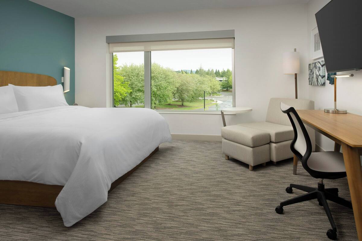 A hotel room with a large bed, a desk with a chair, a small table, and a window with a view of trees and a road.