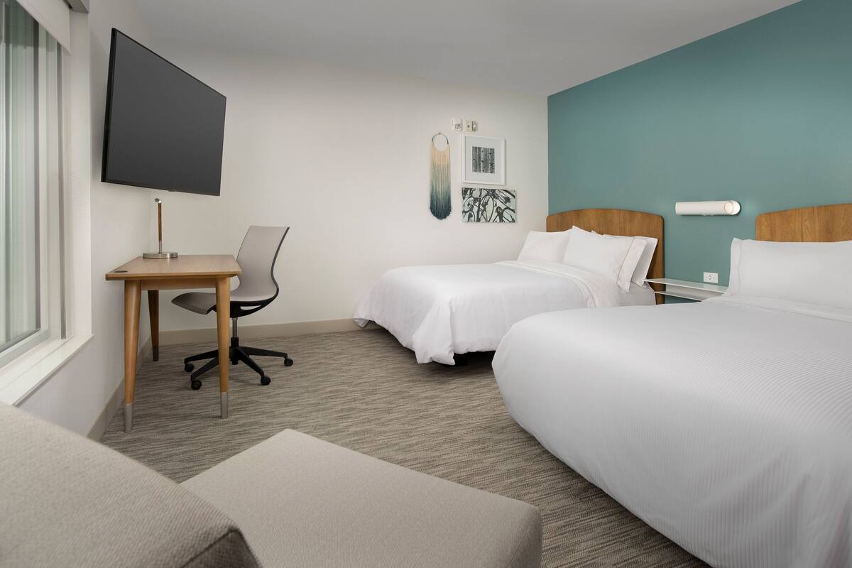 A hotel room with two beds, a desk, a chair, a TV, and artwork on the wall.