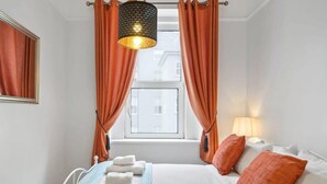 Ein Hotelzimmer mit Bett, orangefarbenen Vorhängen, einer Hängelampe und einem Fenster mit Blick auf Gebäude.