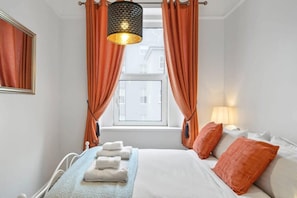 Habitación de hotel con una cama, cortinas naranjas, una lámpara colgante y una ventana que ofrece vistas a edificios.
