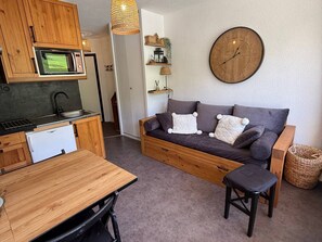 Een knus woongedeelte met een kitchenette, een bank, een eettafel en een wandklok.