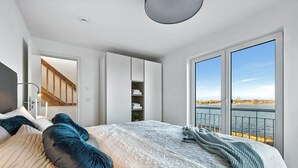 A bedroom with a large bed, a wardrobe, a built-in shelf, and a view of a body of water through a sliding door. - Lütthoorn-1st row by the water, wood-burning stove, sauna, roof terrace, barbecu (Olpenitz)