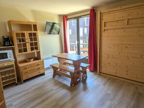 Um quarto com uma mesa de madeira e bancos, micro-ondas, armário com portas de vidro e televisão na parede.