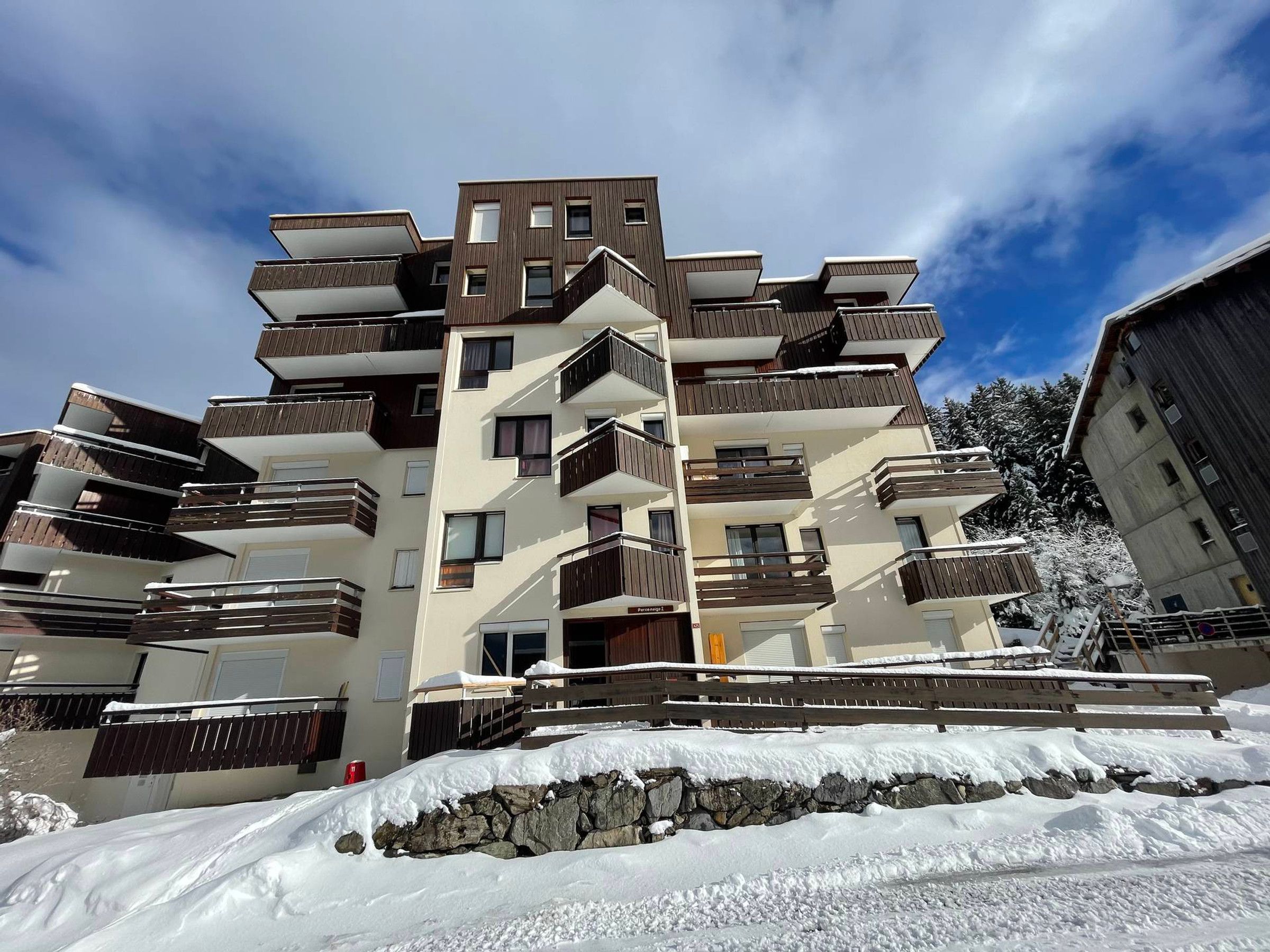 A multi-story residential building with balconies, a stone retaining wall, and snow on the ground.
