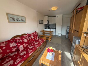 A cozy living room with a red patterned sofa, a wooden coffee table with a vase, and a kitchen area in the background.