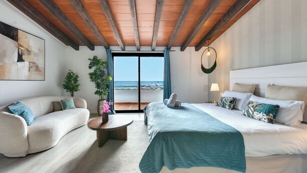 Een slaapkamer met een groot bed, een slaapbank, een klein tafeltje en uitzicht op zee door een schuifdeur.