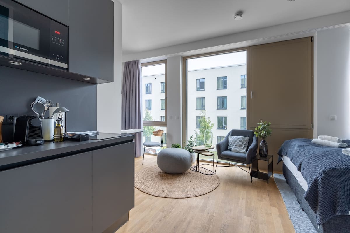 Una habitación de hotel moderna con kitchenette, un ventanal con vista a edificios, una cama confortable y un área de estar con un reposapiés redondo.