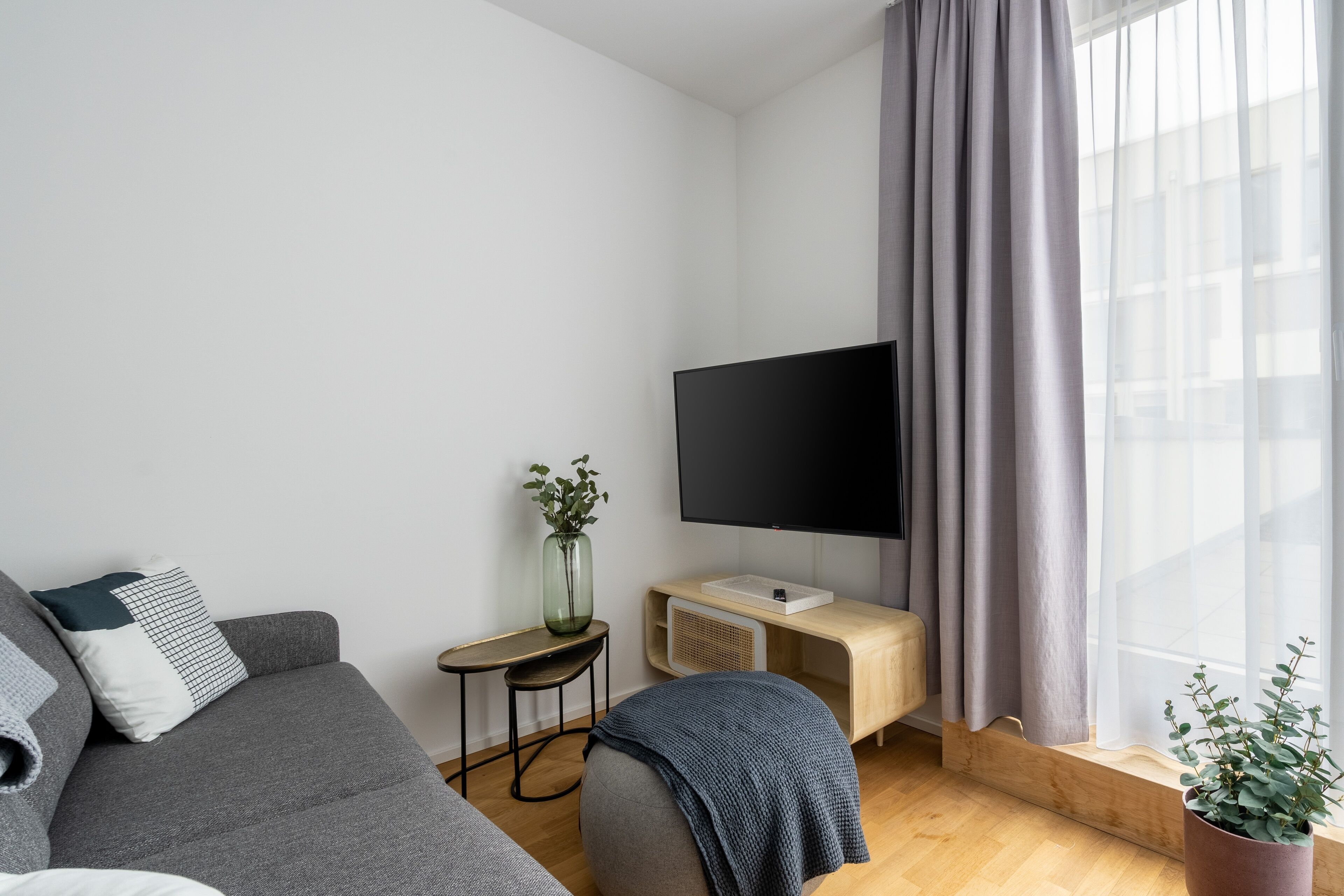 Ein modernes Wohnzimmer mit einem grauen Sofa, einem Flachbildfernseher, einem kleinen Beistelltisch mit einer Vase und einem Fenster mit grauen Vorhängen.