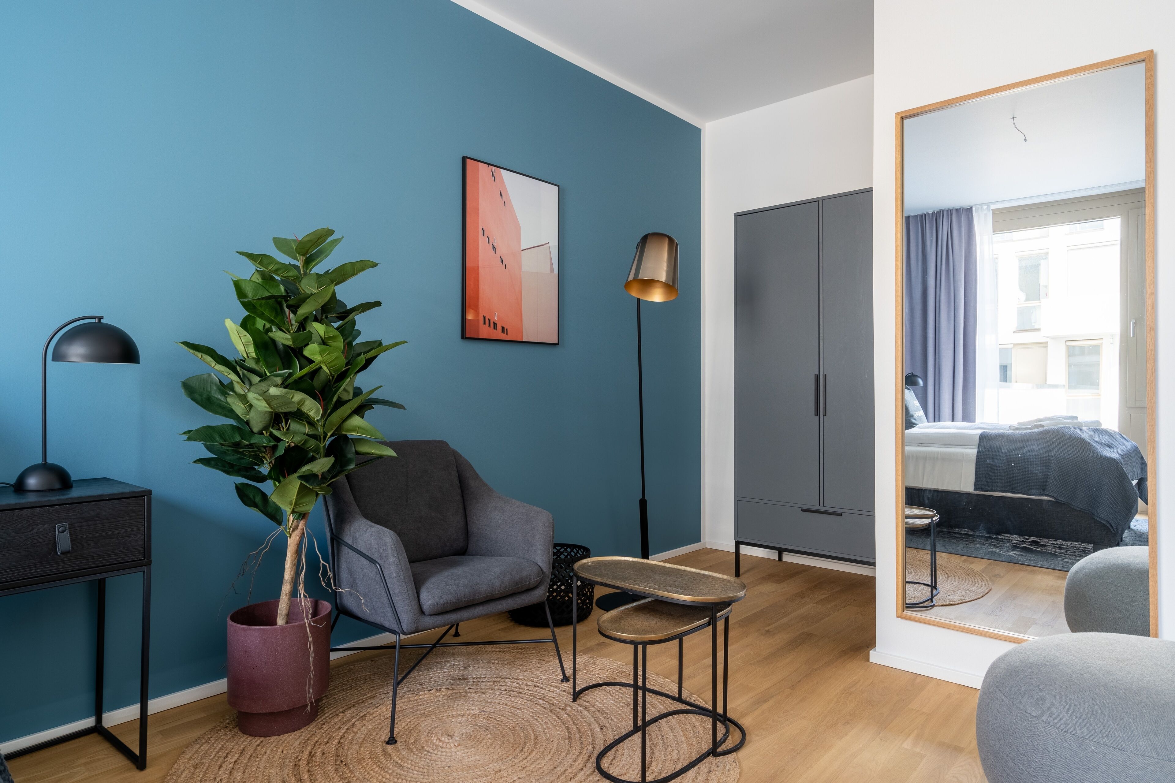 Een moderne woonkamer met een blauwe accentmuur, een grijze fauteuil, een houten salontafel, een vloerlamp en een grote spiegel die een slaapkamer met een bed en gordijnen reflecteert.