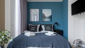 A neatly made bed with two pillows labeled "SUITE DREAMS" and "SUITE MORNING," a blue wall, a mounted TV, and a floor fan.