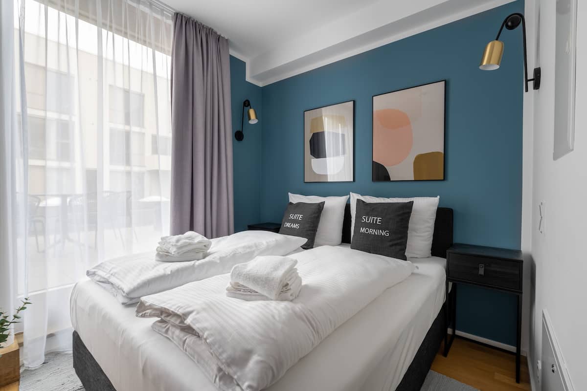 Un dormitorio moderno con una cama grande, dos mesitas de noche y vistas a la ciudad a través de cortinas traslúcidas.