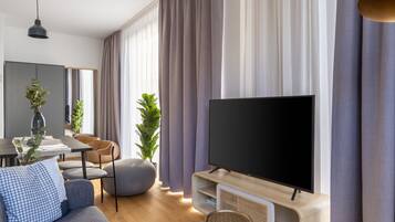 Una sala moderna con un televisor de pantalla plana, sofá gris, mesa de comedor y ventanas grandes con cortinas.