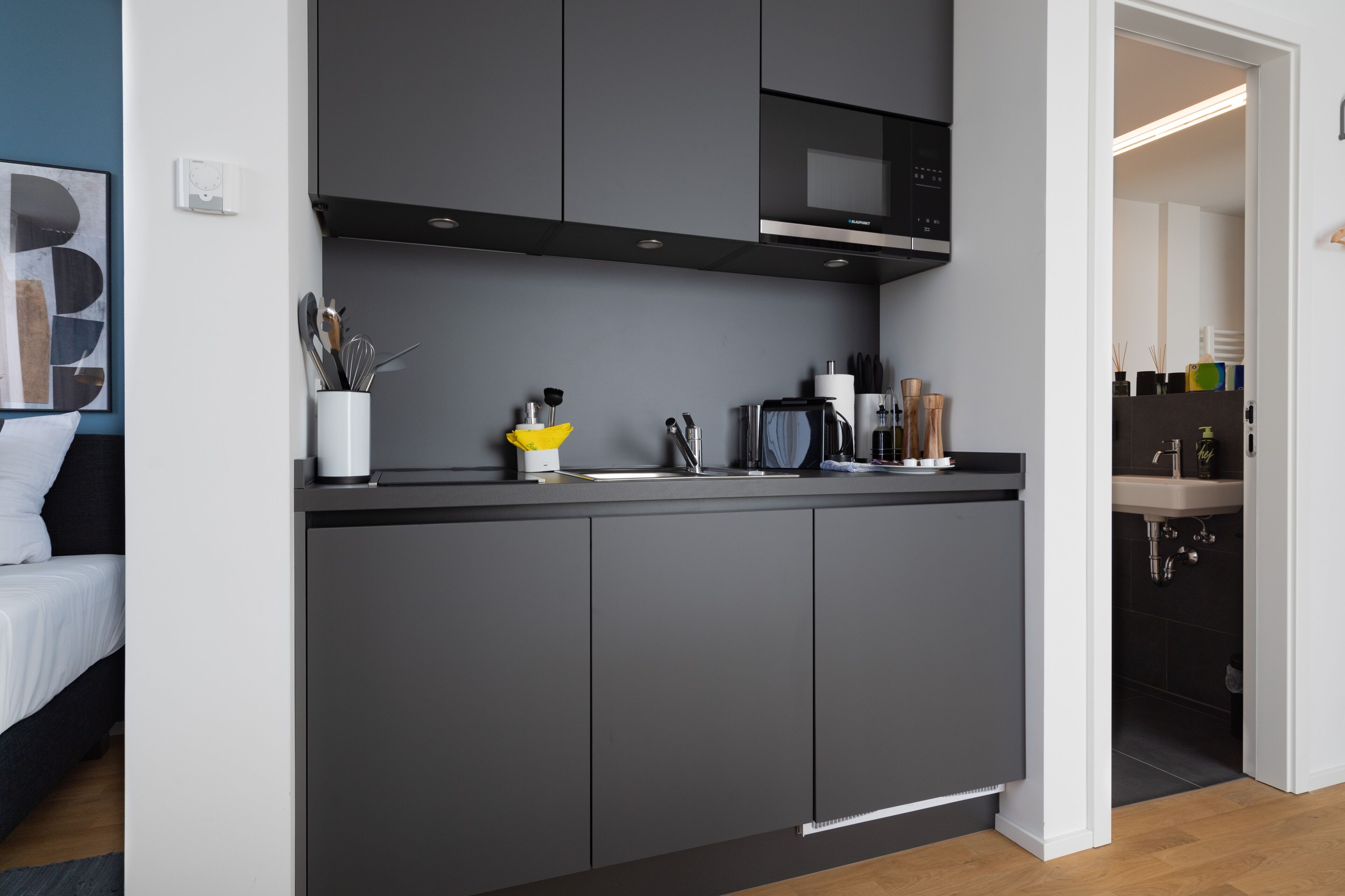 Una cocina moderna con armarios grises, un microondas y un lavamanos. En la habitación contigua hay una cama con ropa de cama blanca.