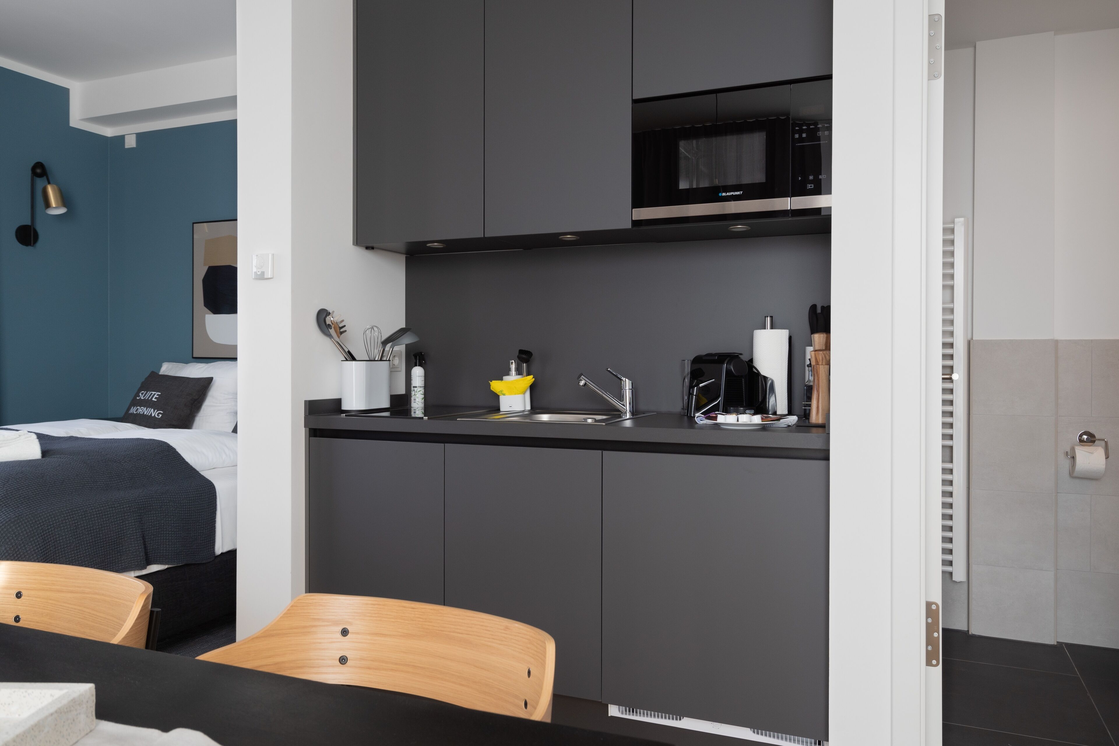 Habitació d'hotel moderna amb petita cuina, un llit amb capçaleri blau fosc, una cadira de fusta i una taula petita.