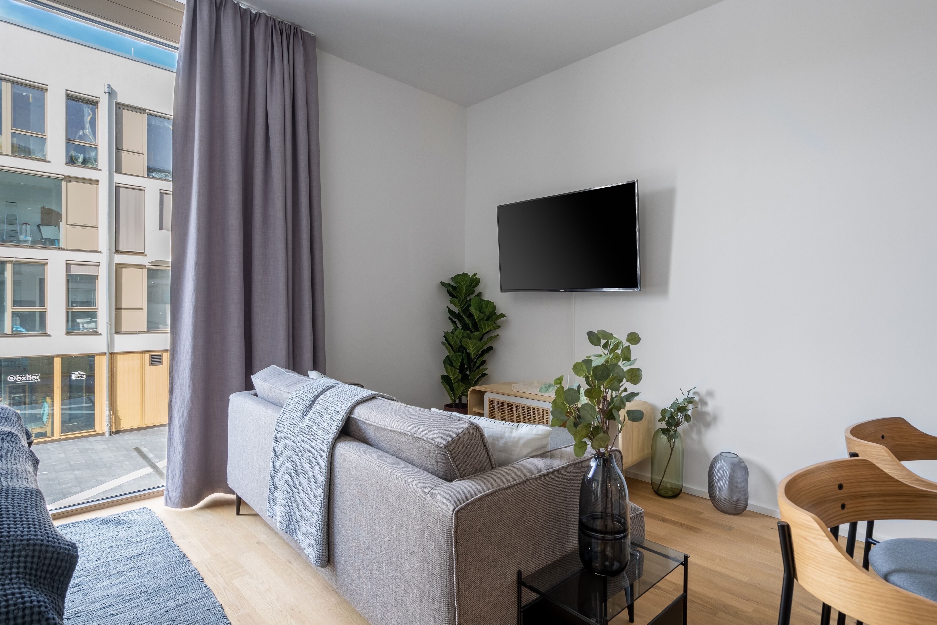 A modern living room with a grey sofa, a flat-screen TV mounted on the wall, a small table with a vase, and a view of a building outside.