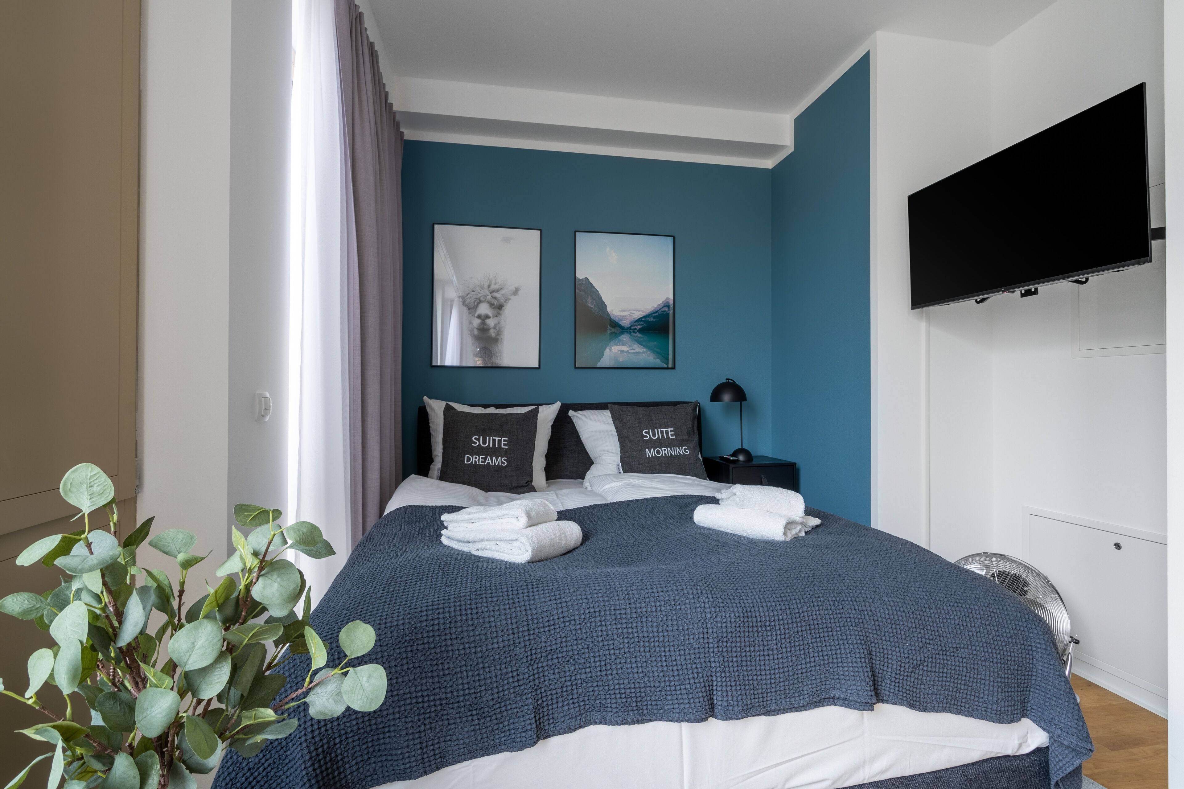 A neatly made bed with a blue and white duvet, two pillows with text, a bedside table with a lamp, and a wall-mounted TV.