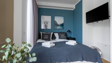 A neatly made bed with a blue and white duvet, two pillows with text, a bedside table with a lamp, and a wall-mounted TV.