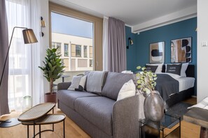 A modern living room with a grey sofa, a coffee table, a potted plant, and a bed with a dark headboard. - Limehome Potsdam Annemarie-Wolff Platz (Potsdam)