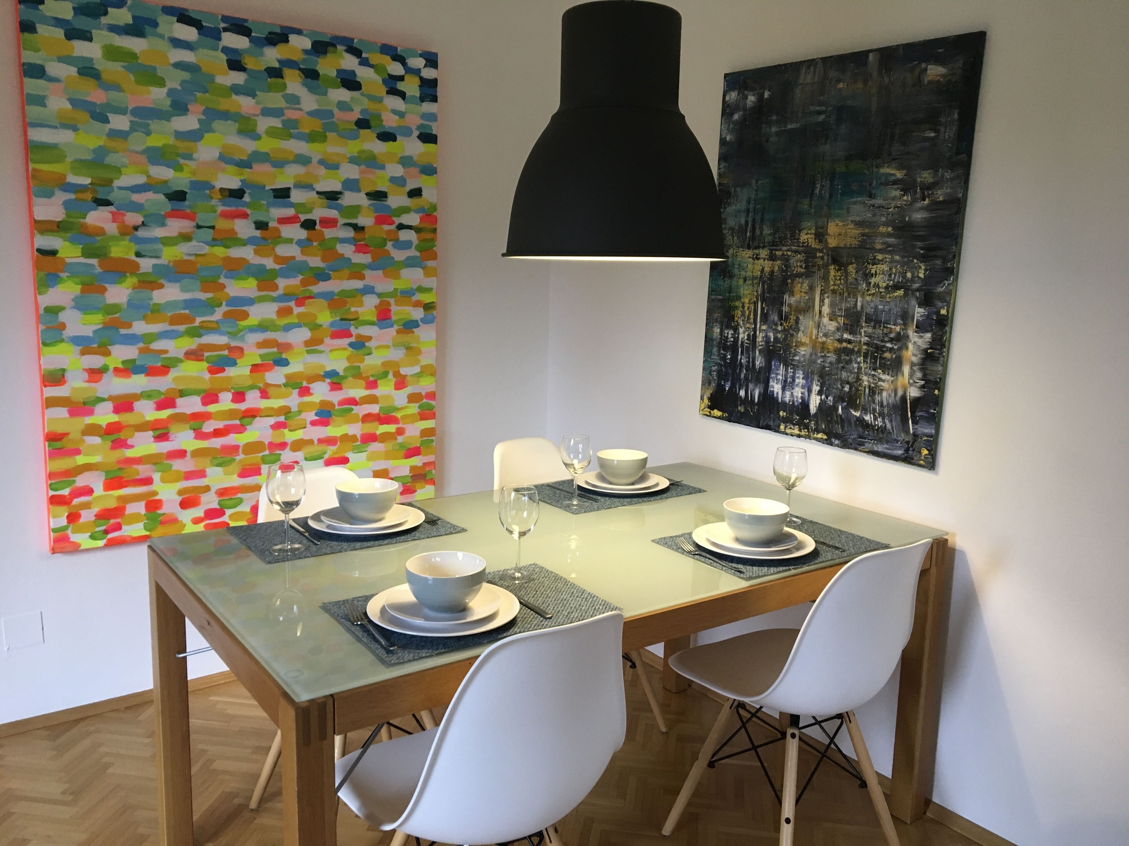 Zona pranzo con tavolo in vetro, sedie bianche e lampadario nero a sospensione. Sul tavolo ci sono due ciotole e due bicchieri da vino.