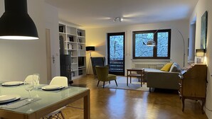 Un salón moderno con comedor, piso de madera y ventanas amplias con vistas a árboles.