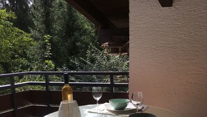 Una mesa redonda de vidrio con mantel blanco, una botella y un bol sobre un balcón con vistas a árboles y barandilla de madera.