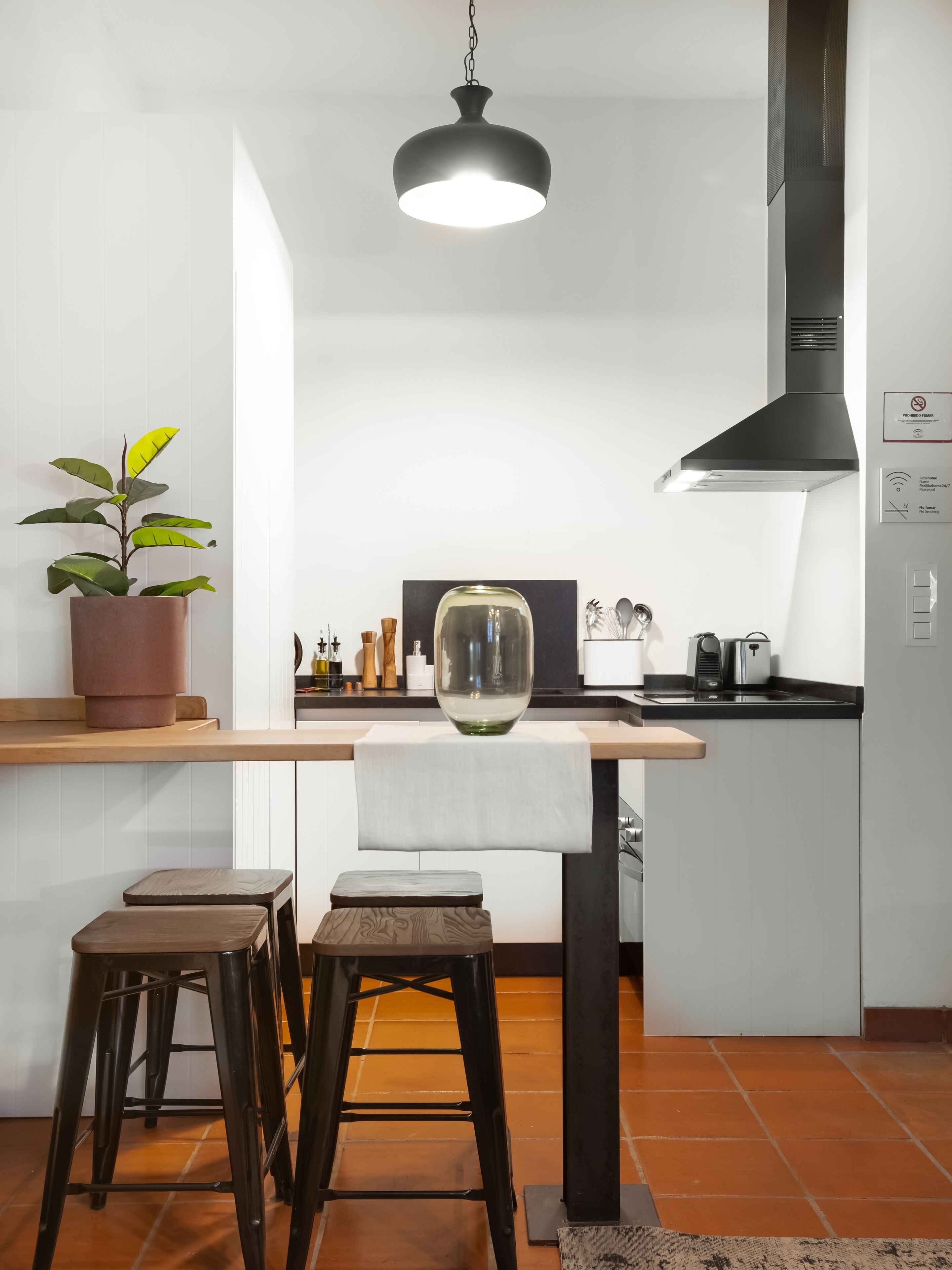 A modern kitchen with a central island, stools, a hanging light fixture, and a potted plant.