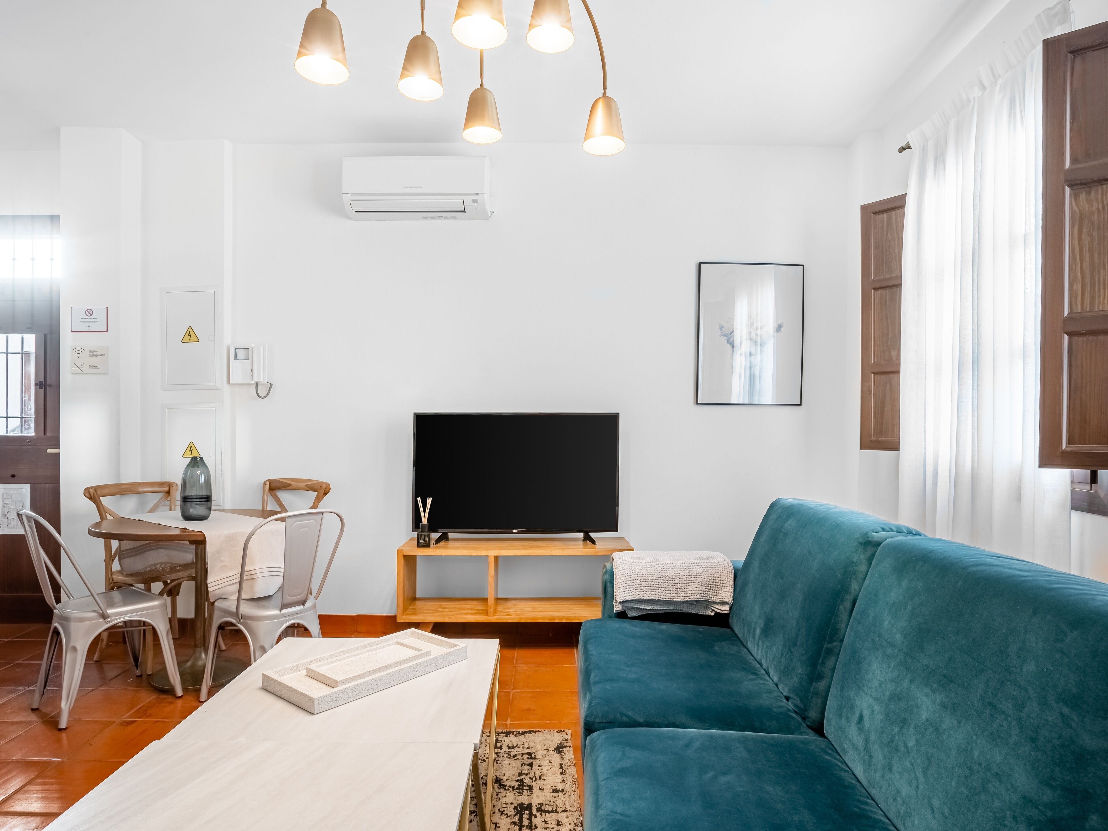 A modern living room with a teal sofa, a wooden dining table with chairs, a flat-screen TV, and pendant lights.