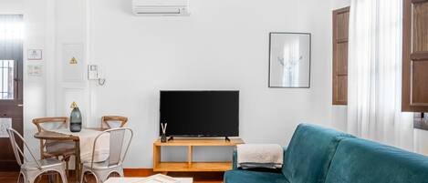 A modern living room with a teal sofa, a wooden dining table with chairs, a flat-screen TV, and pendant lights.