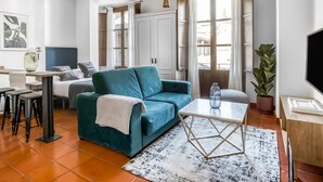 Ein modernes Wohnzimmer mit einem türkisfarbenen Sofa, einem Couchtisch und einem Fernseher. Es gibt einen Essbereich mit Tisch und Stühlen sowie eine Küchenzeile mit Schränken und einer Pflanze.