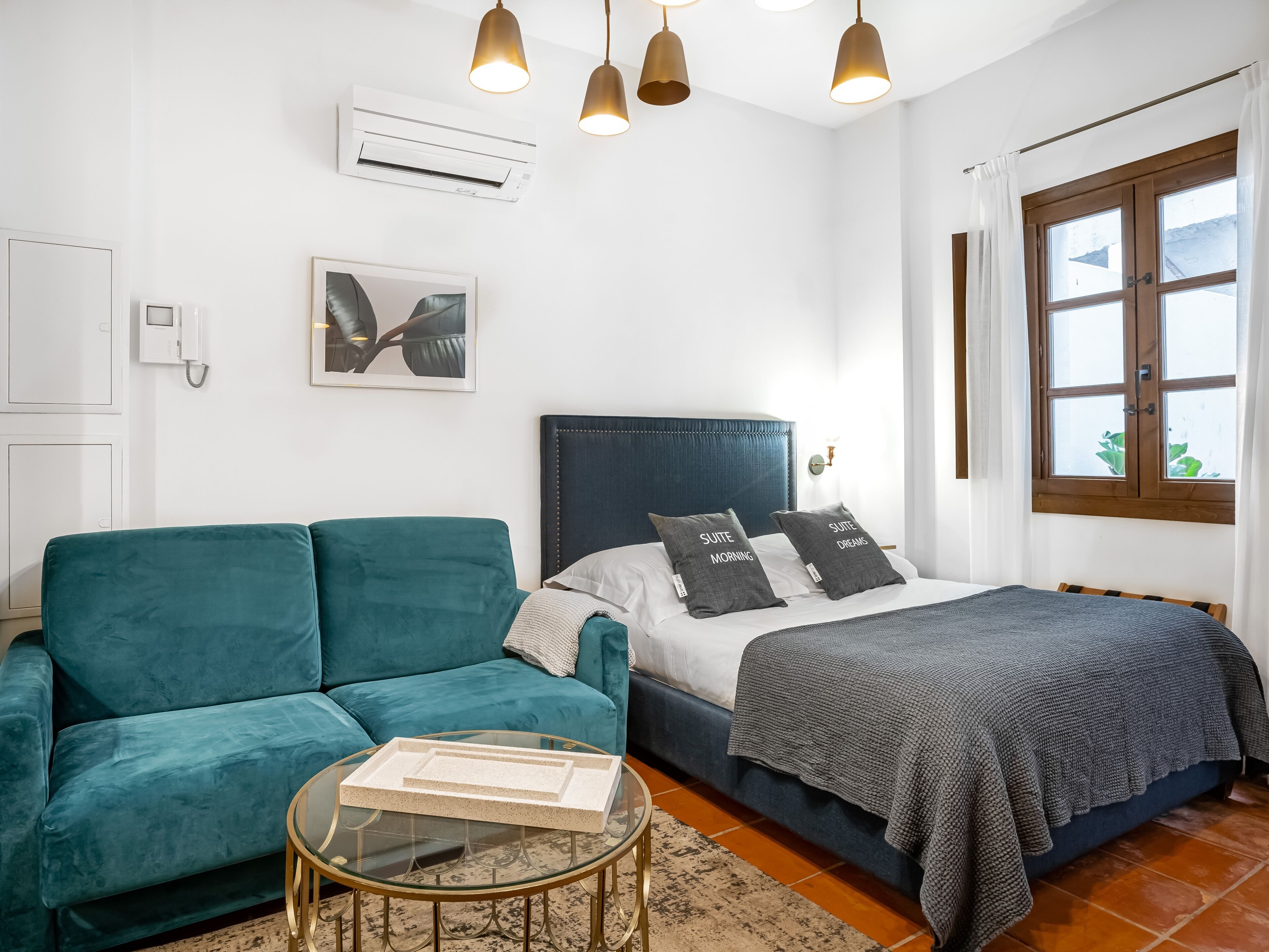 Un dormitorio con cama, un sofá azul, una mesita de centro de cristal y una ventana con cortinas.