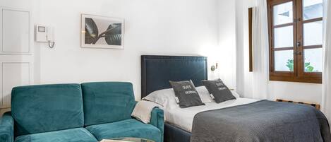 A bedroom with a bed, a blue sofa, a glass coffee table, and a window with curtains.