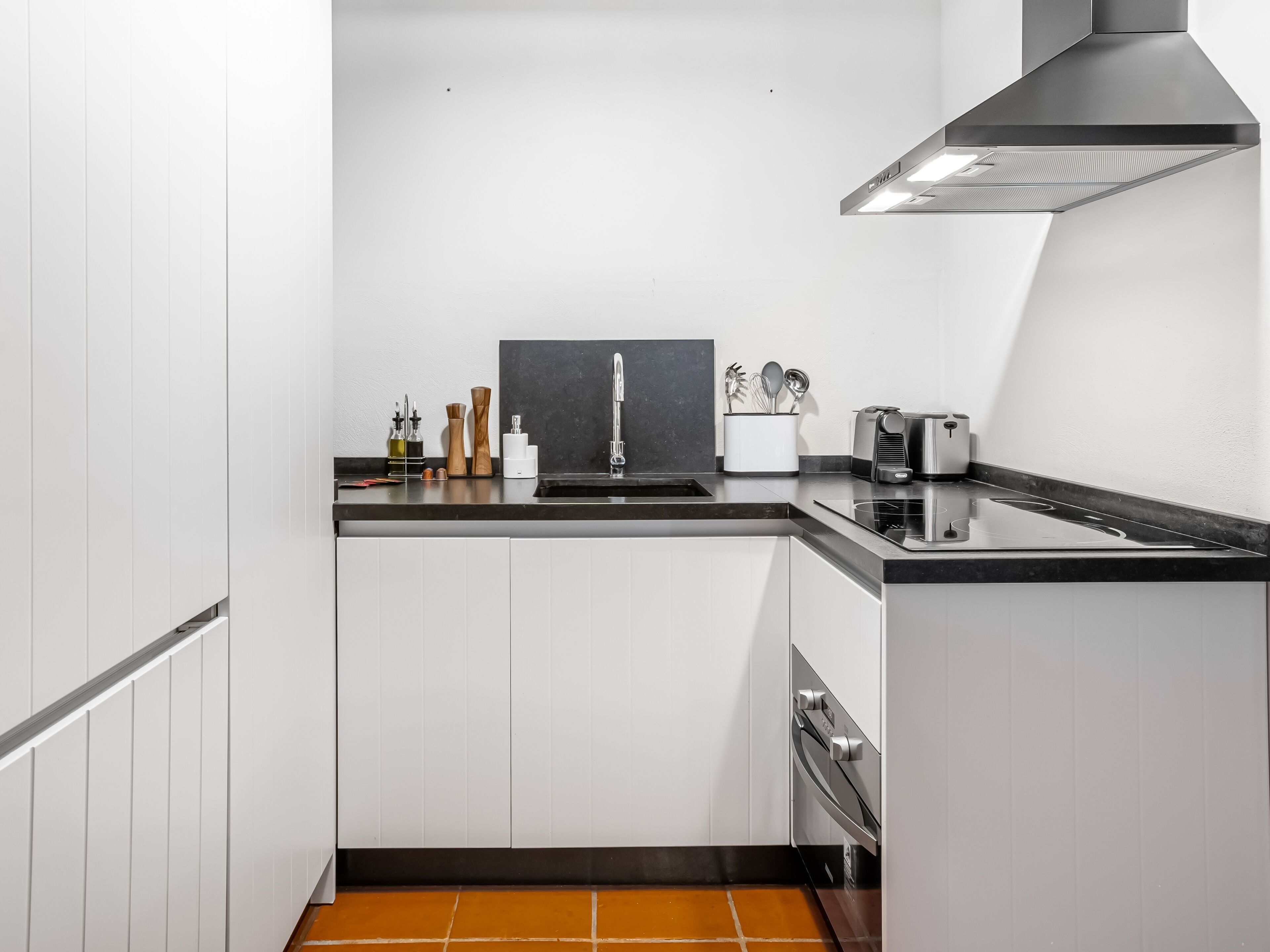 Une cuisine compacte avec des meubles blancs, un plan de travail noir, une hotte en acier inoxydable et un four encastré.