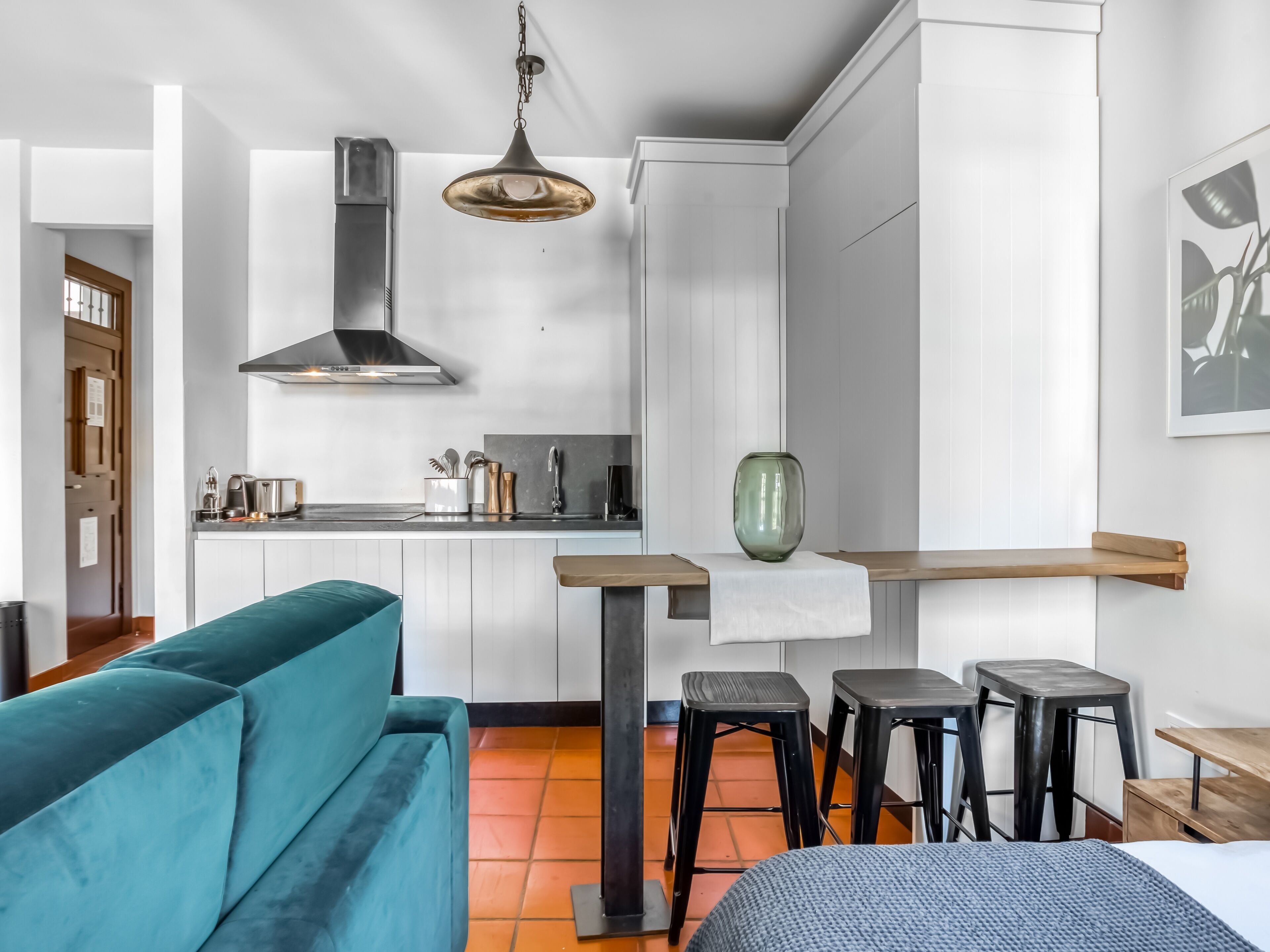 A modern kitchen with a stainless steel range hood, a dining area with a wooden table and black stools, and a teal sofa.