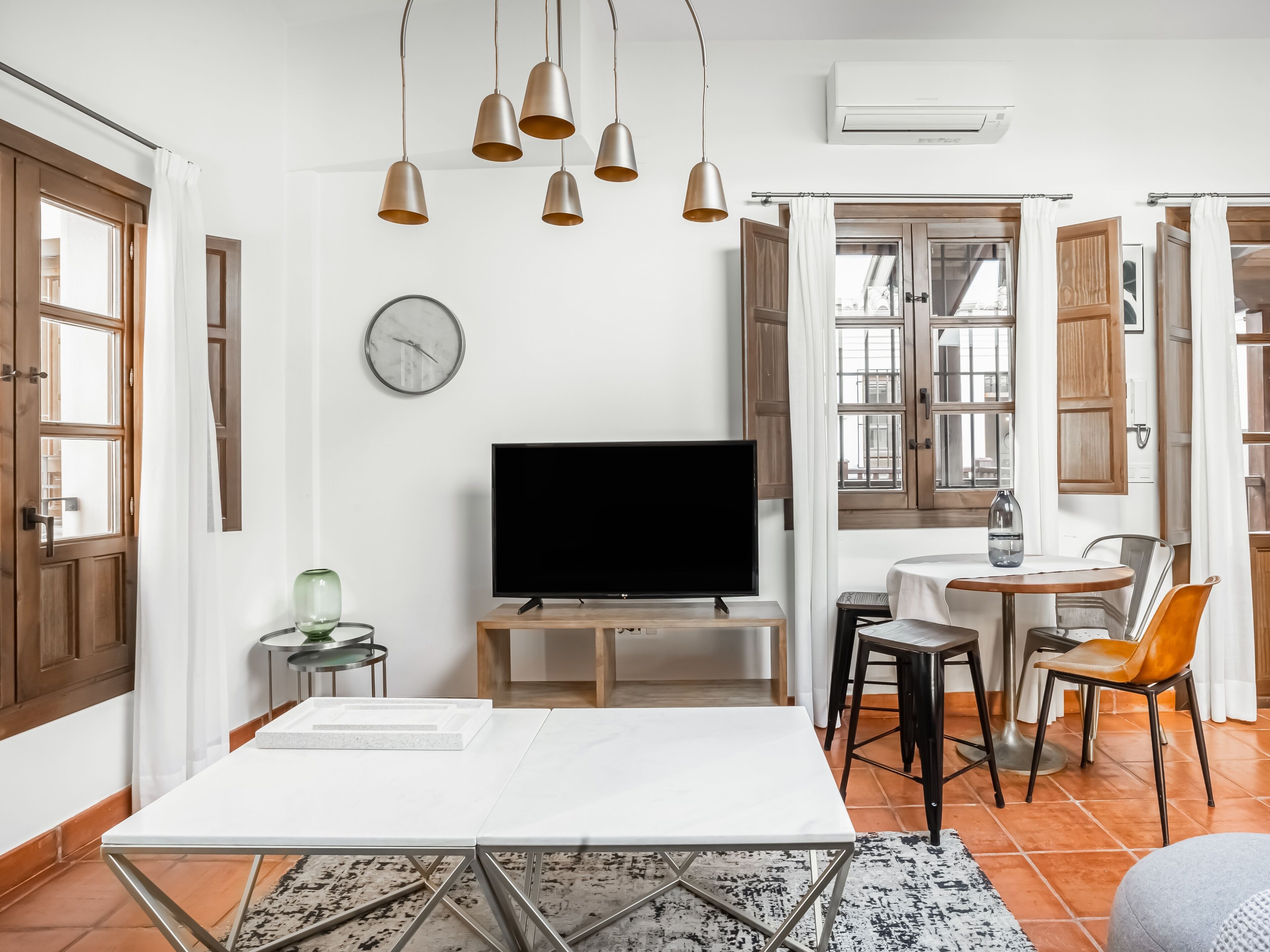 A modern living room with a flat-screen TV, a dining table, and a coffee table. There are pendant lights, a clock, and a window with wooden shutters.