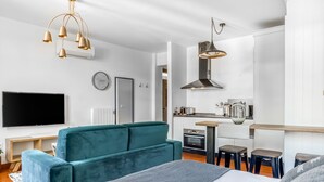 Ein modernes, helles Zimmer mit einem großen Bett, einem türkisblauen Sofa, einem Fernseher und einer Küchenzeile.