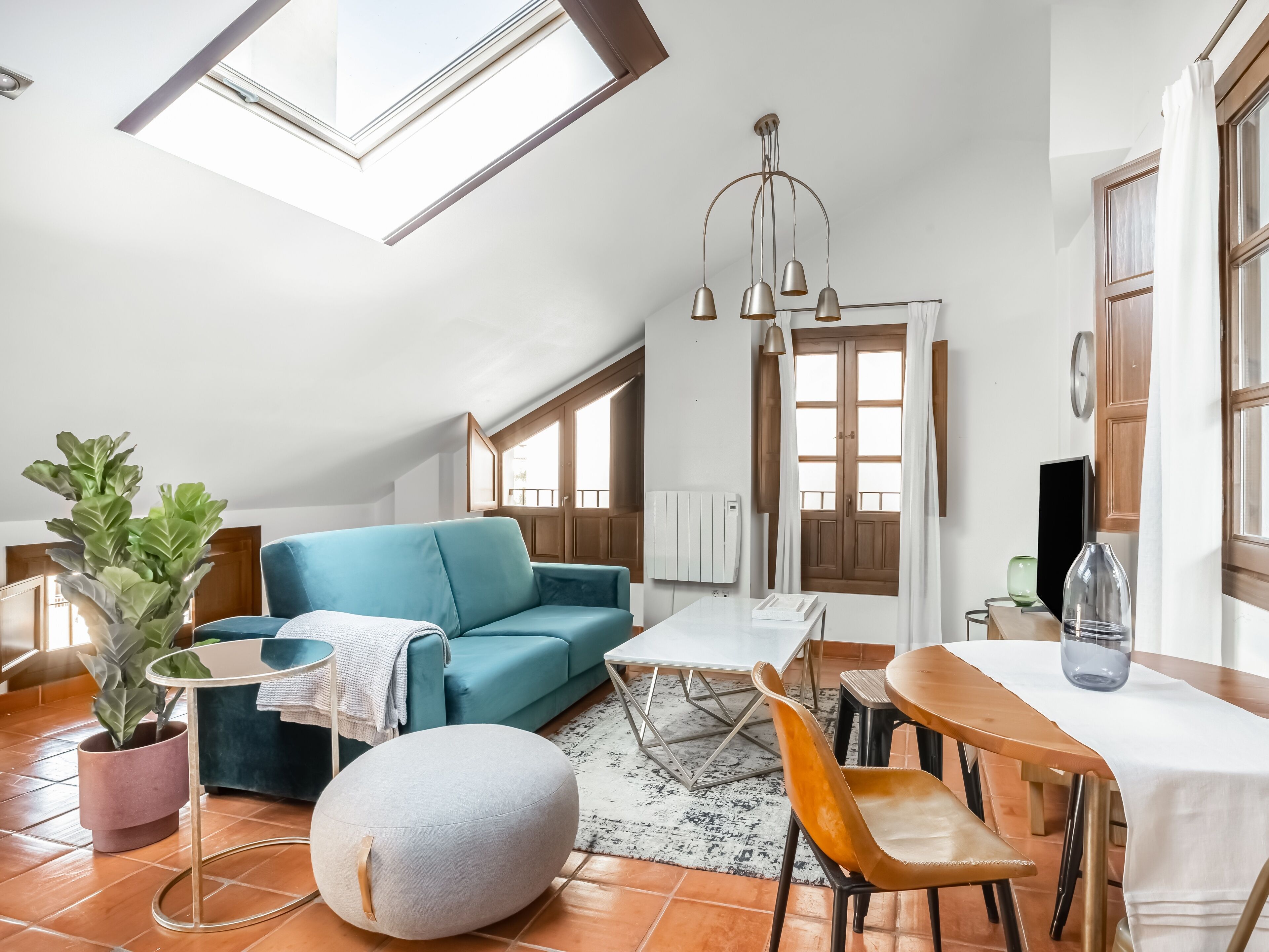 Una sala de estar moderna con un sofá azul, un taburete redondo, una mesa de comedor con sillas, una planta en maceta y un amplio ventanal con cortinas.