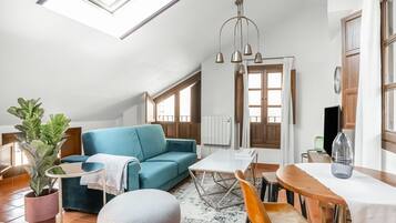 Una sala de estar moderna con un sofá azul, un taburete redondo, una mesa de comedor con sillas, una planta en maceta y un amplio ventanal con cortinas.