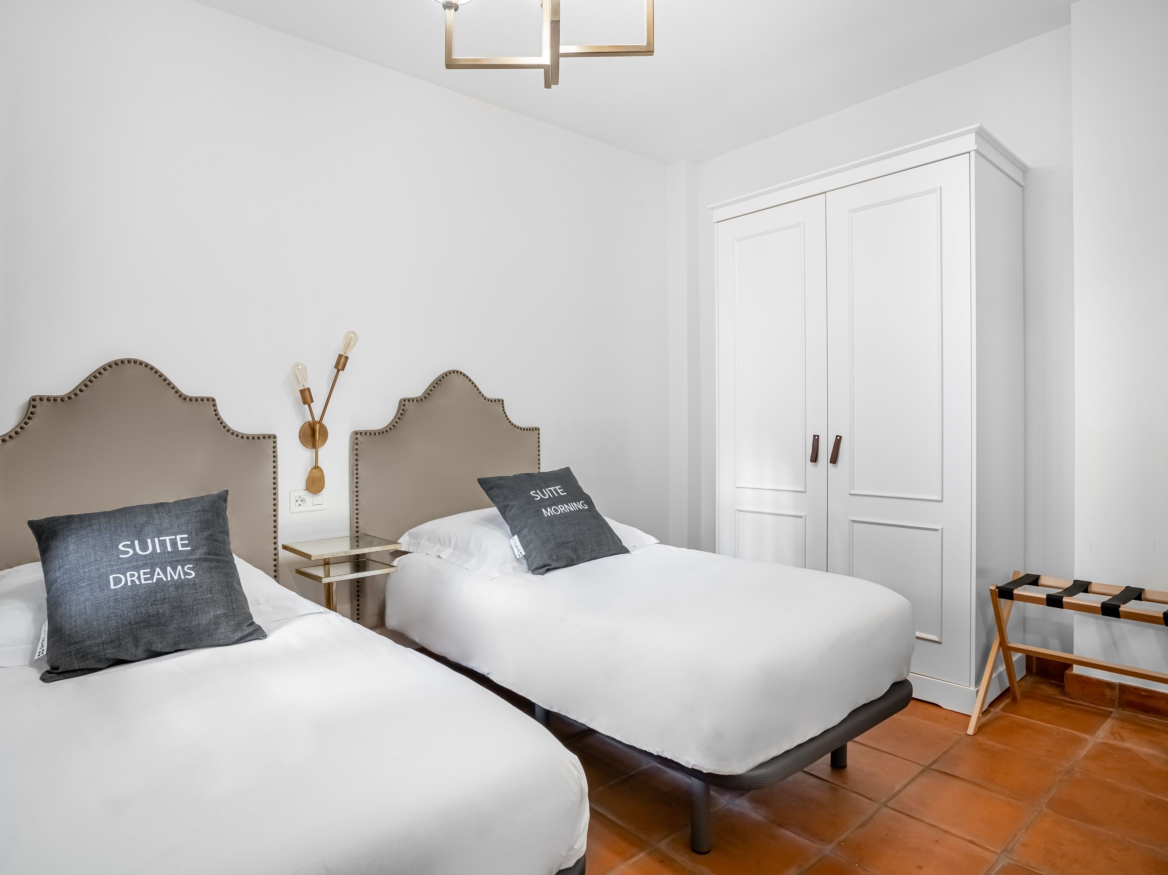 A hotel room with two beds, a wardrobe, a bedside table with a lamp, and a small bench.