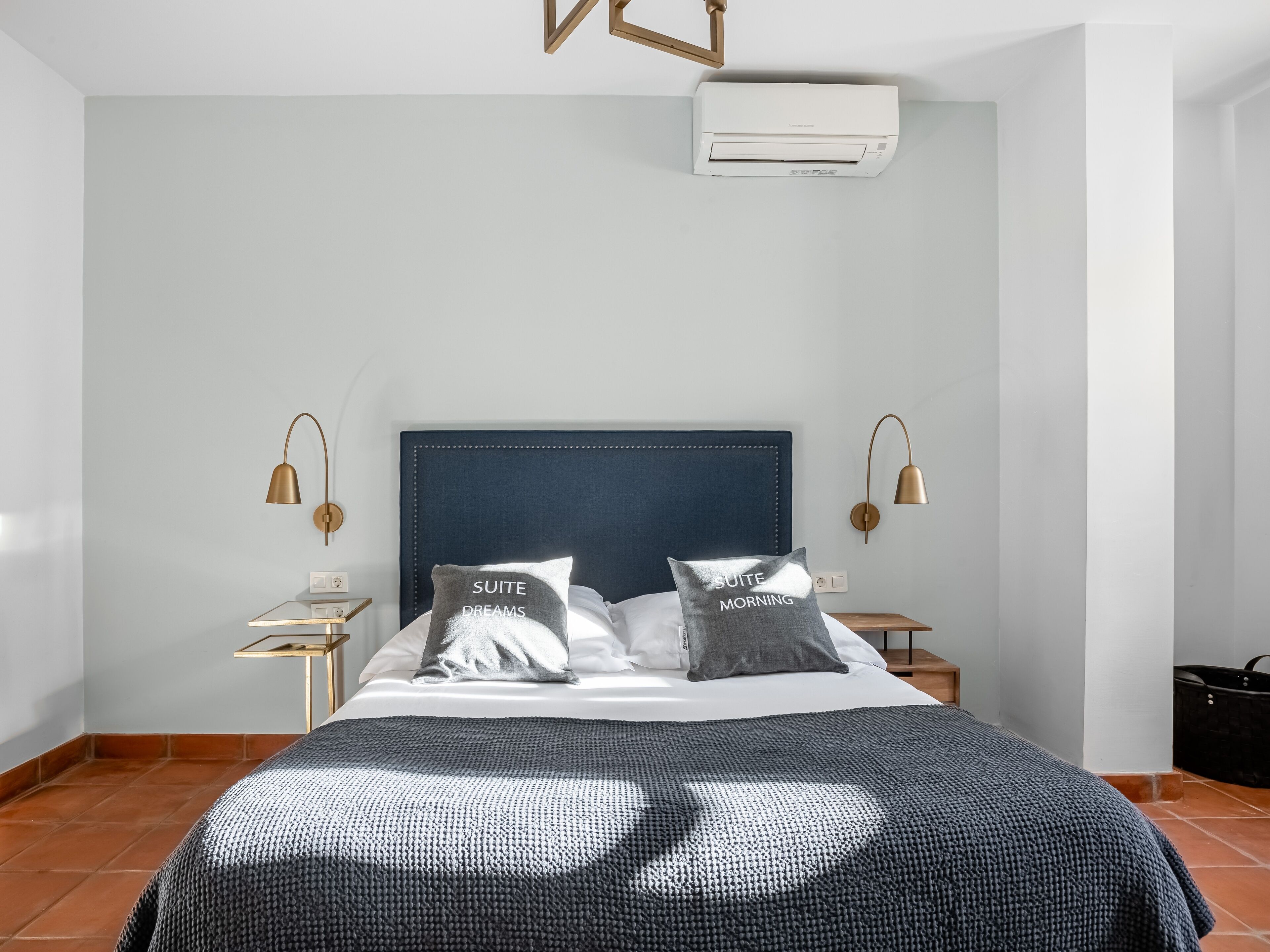 Una habitación de hotel moderna con una cama grande, dos mesitas de noche, un aire acondicionado de pared y una pequeña mesita auxiliar con una lámpara.