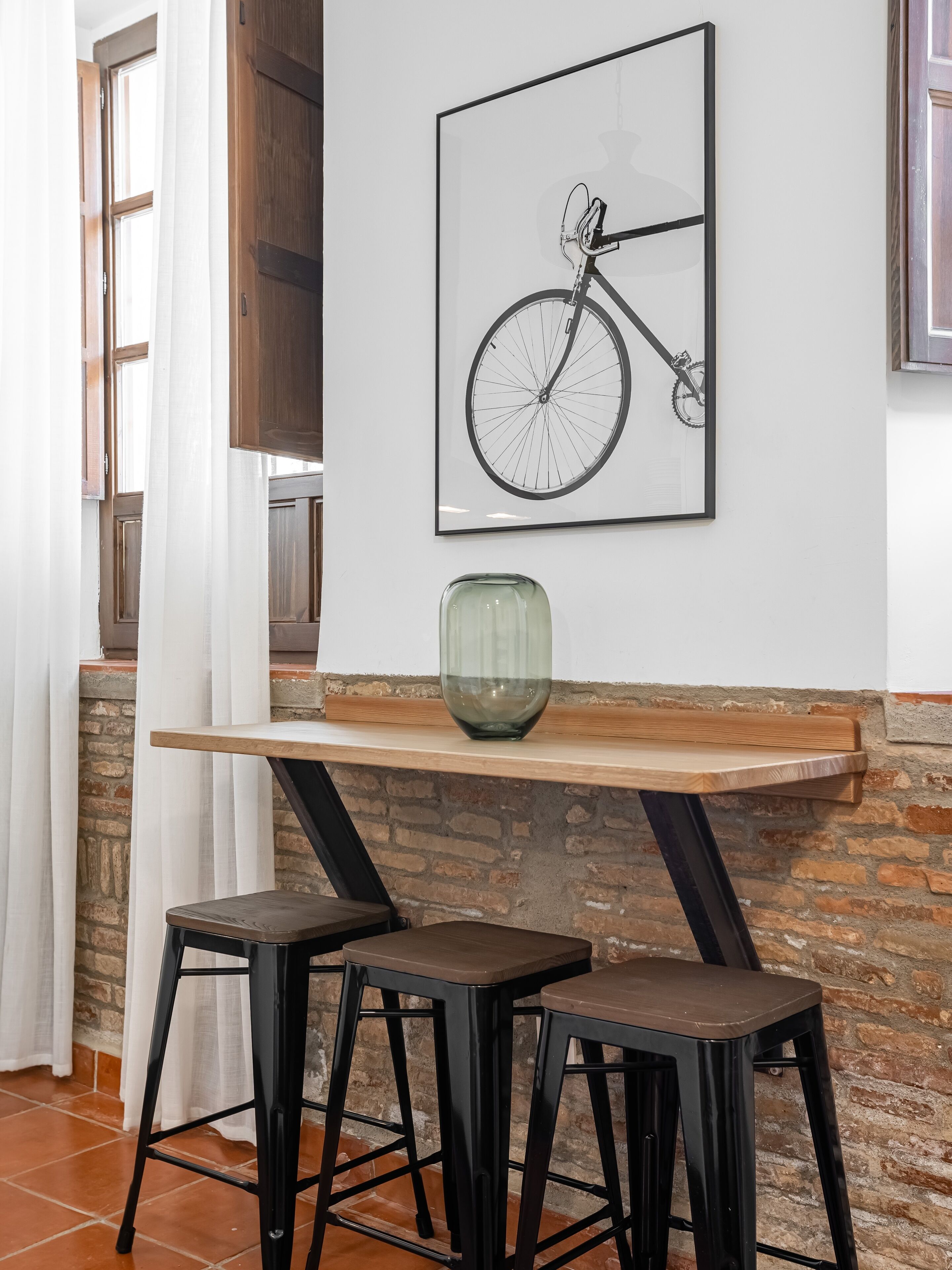 A dining area with a wooden table, two black stools, a framed bicycle artwork, and a green vase.